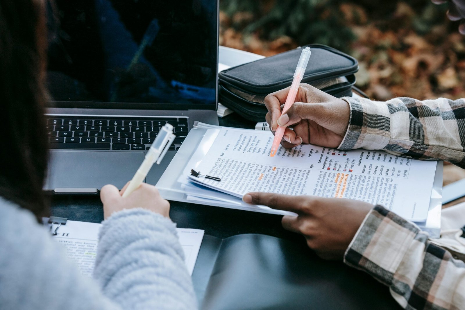High angle of crop anonymous diverse female students highlighting information in printed texts in papers while sitting at table with laptop in campus
