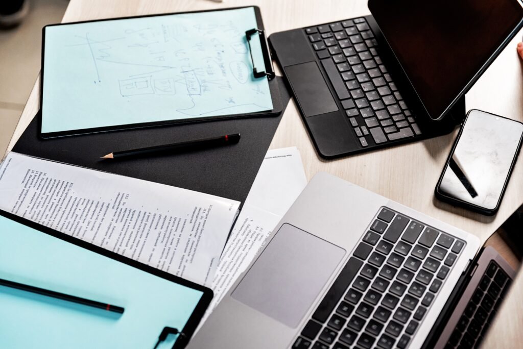 Workspace scene with two laptops, a clipboard on blue paper, a pencil, a tablet, and scattered documents on a wooden desk